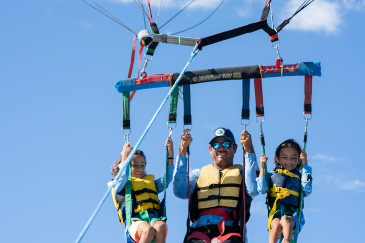 Three people parasailing with a blue parachute against a clear sky.