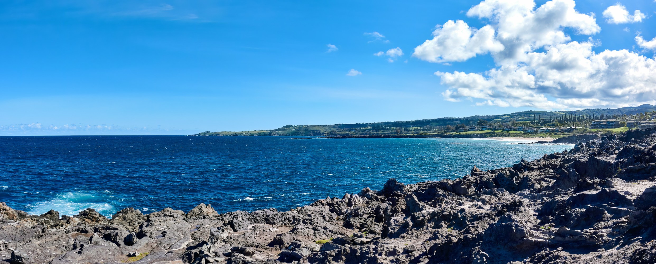Rocky coastline overlooking blue ocean under a clear sky with scattered clouds.
