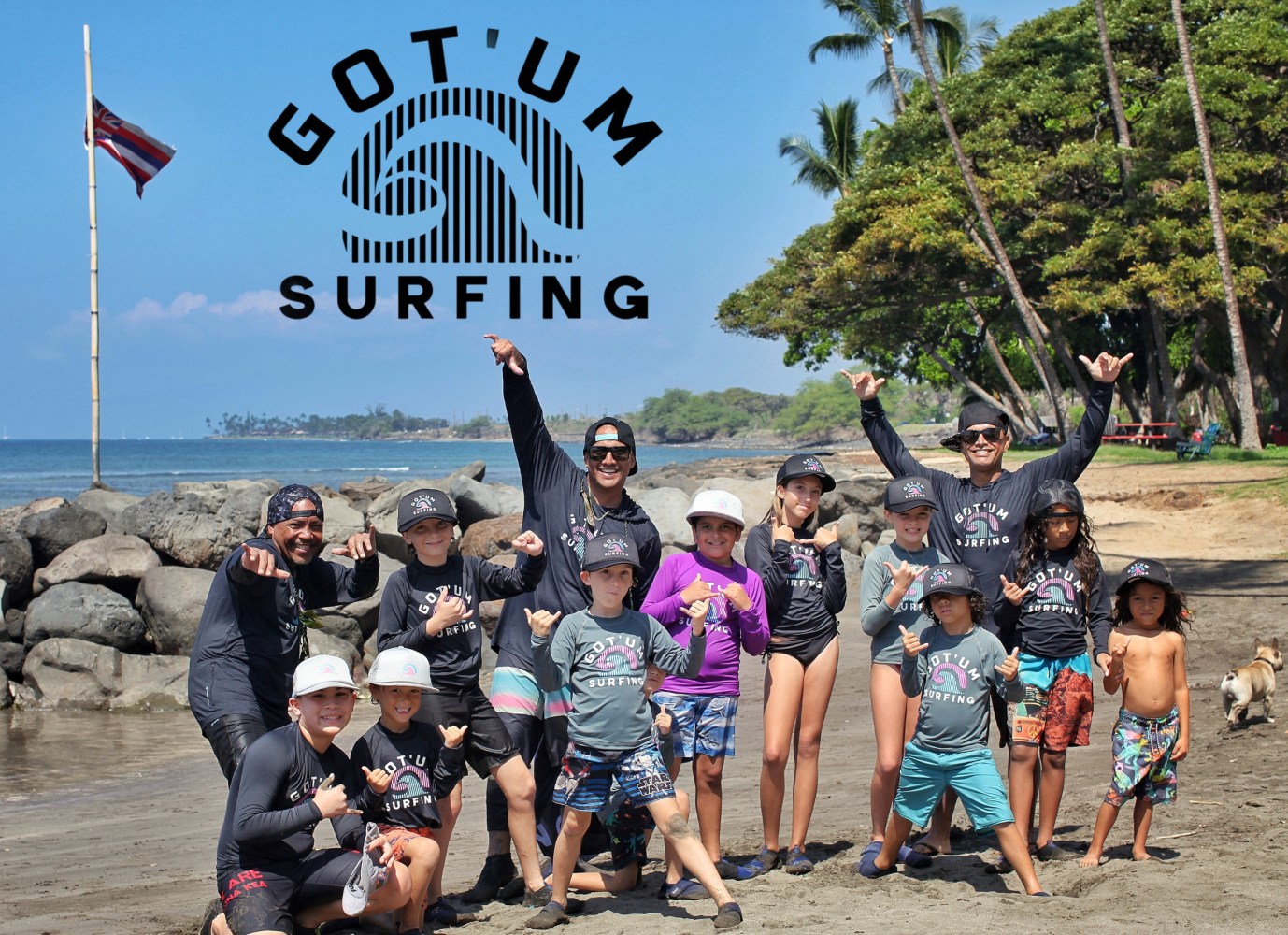 Group of children and adults on a beach posing with hand signs, wearing 'Got'um Surfing' shirts and caps.