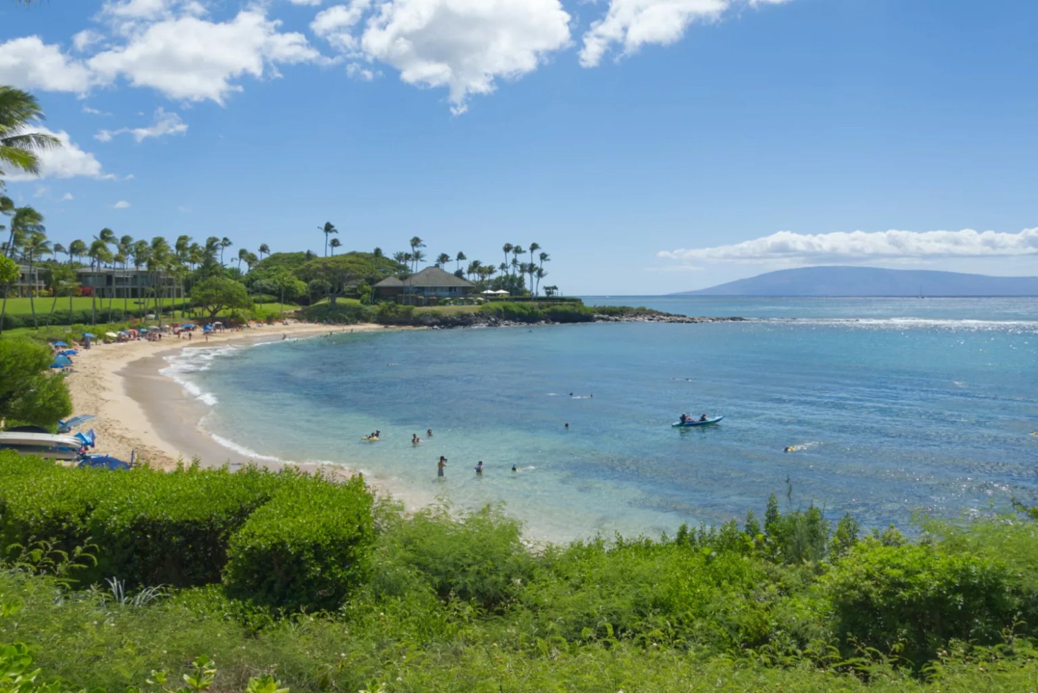 A sandy beach cove with clear blue water, people swimming, and lush greenery under a sunny sky.