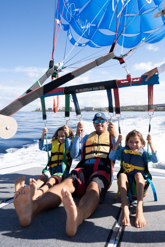 Three people parasailing over water, wearing life jackets, with blue parachute above.