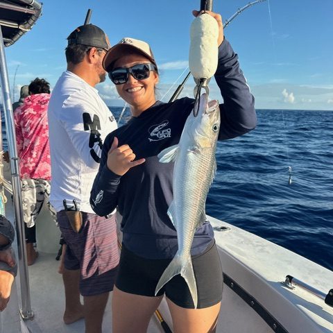 Person holding a fish on a boat, ocean in the background, with another person fishing nearby.