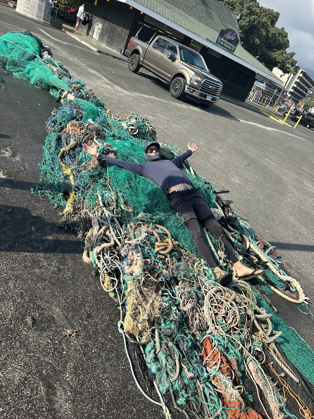 Person lying on large tangled fishing nets in a parking lot with a truck nearby.
