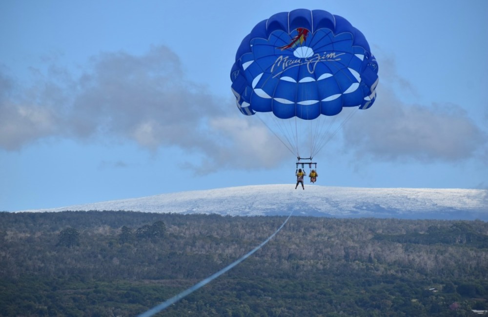 Two people parasailing with a blue parachute over a scenic landscape.