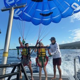 Two people parasailing with a blue parachute, guided by a person on a boat.