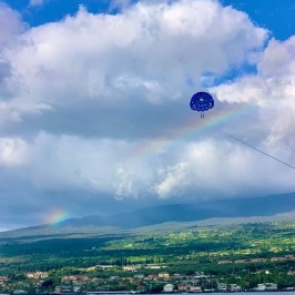 Parasailer under a rainbow above a coastal town with cloudy sky.