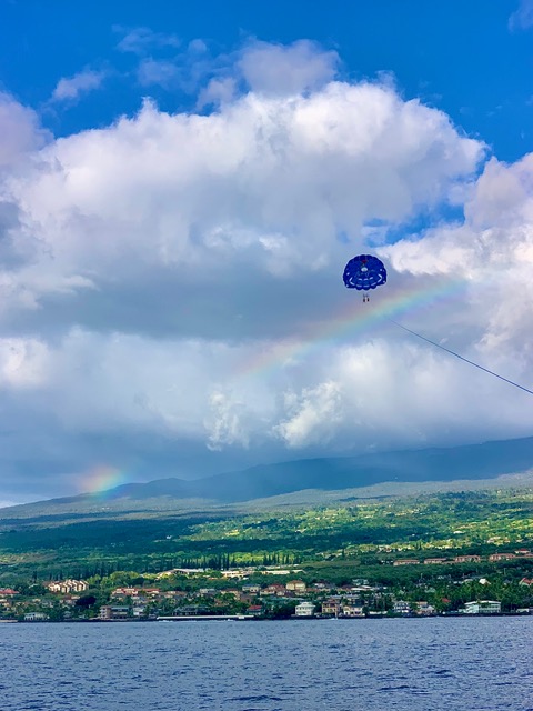 Parasailer under a rainbow above a coastal town with cloudy sky.