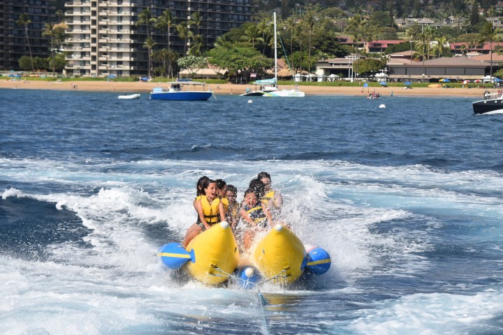 People riding a banana boat on the ocean near a sandy beach with buildings in the background.