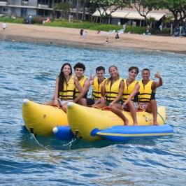 Group of six people on a yellow banana boat in the ocean near the beach.