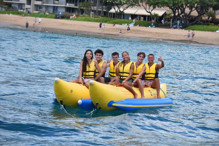 Group of six people on a yellow banana boat in the ocean near the beach.