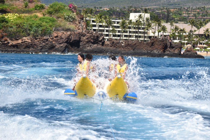 People riding a banana boat on choppy water near a rocky coastline with a hotel in the background.