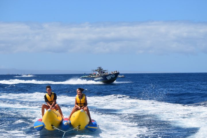 Two people in yellow vests riding a banana boat on the ocean with a speedboat in the background.