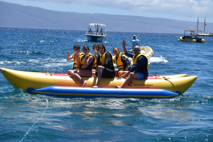 Group of people on a yellow inflatable banana boat in the ocean, waving and wearing life jackets.