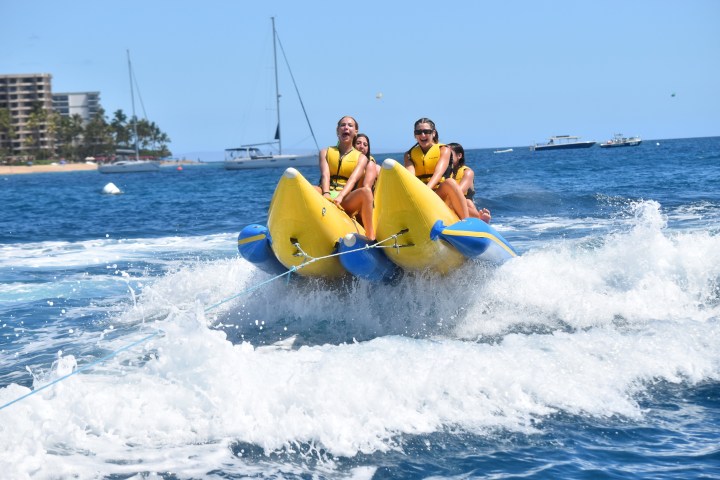Four people on a banana boat ride in the ocean with waves, wearing life jackets.