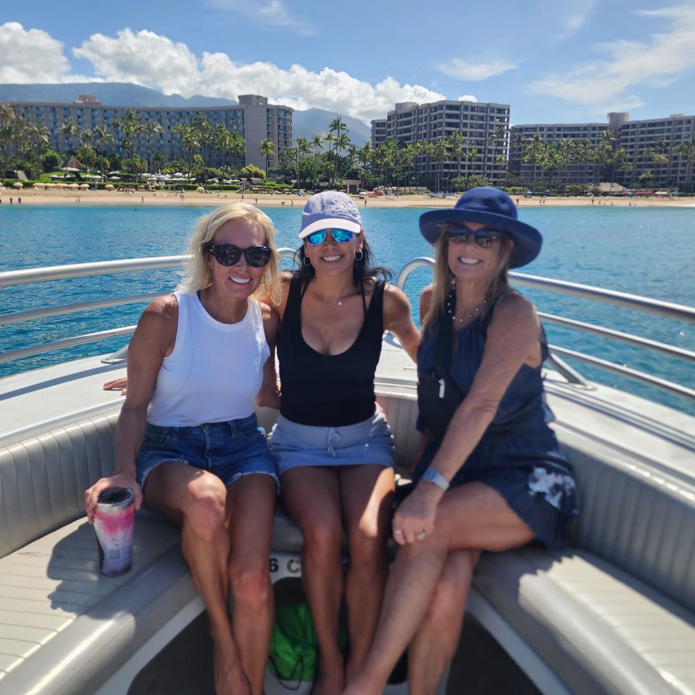Three women sitting on a boat near a beach with buildings in the background.