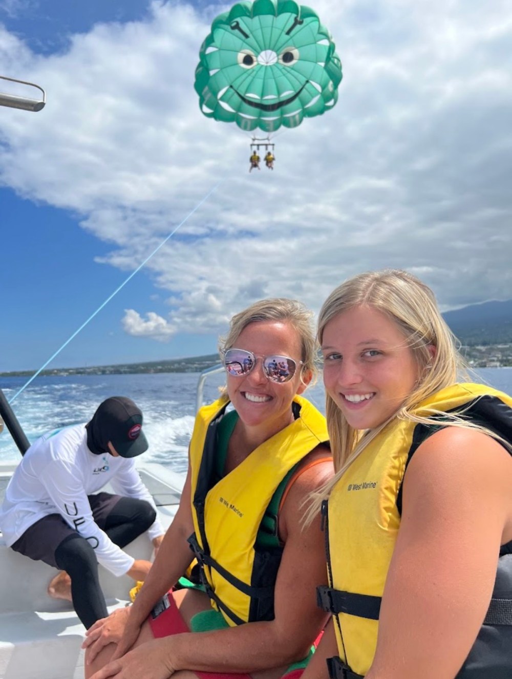 Two women in life jackets on a boat, parasailing with a green parachute in the background.