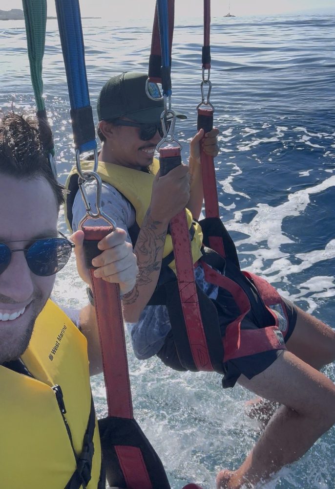 Two people parasailing over the ocean, wearing life jackets and harnesses.