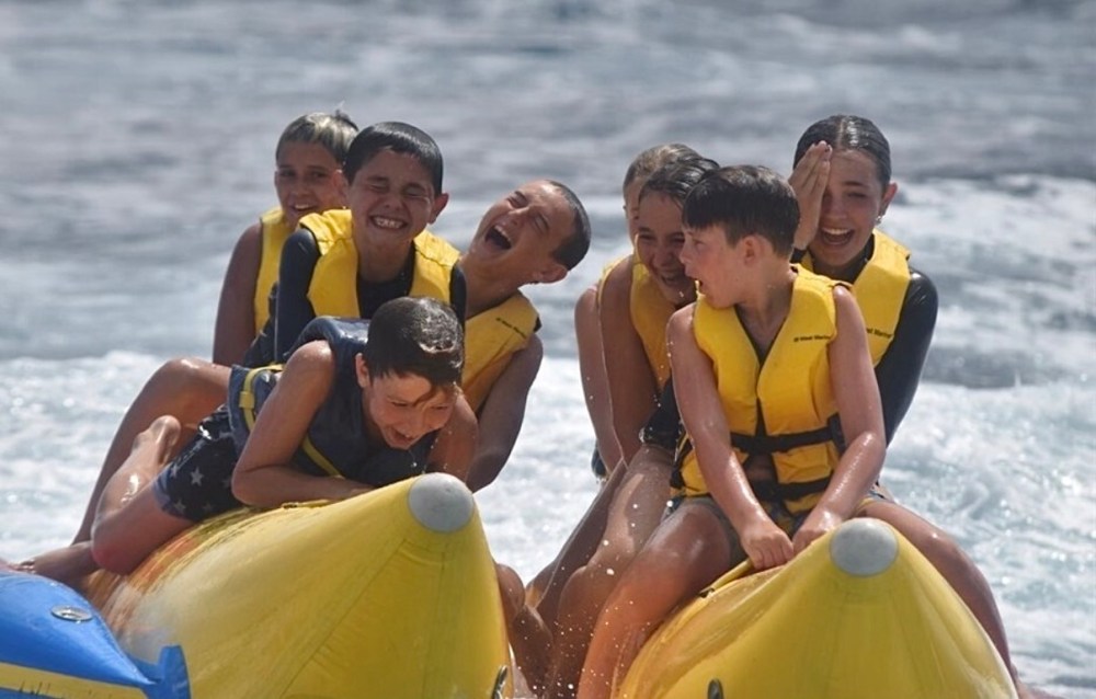 Group of kids in yellow vests laughing on a banana boat in the water.