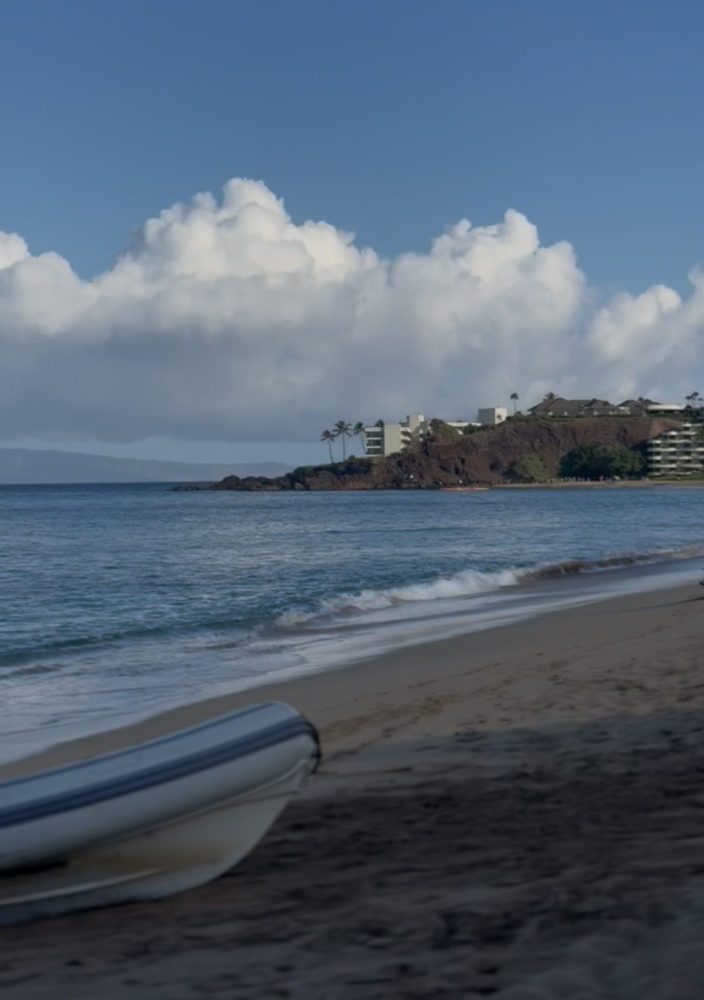 Calm beach with inflatable boat, distant buildings on a hill under cloudy sky.